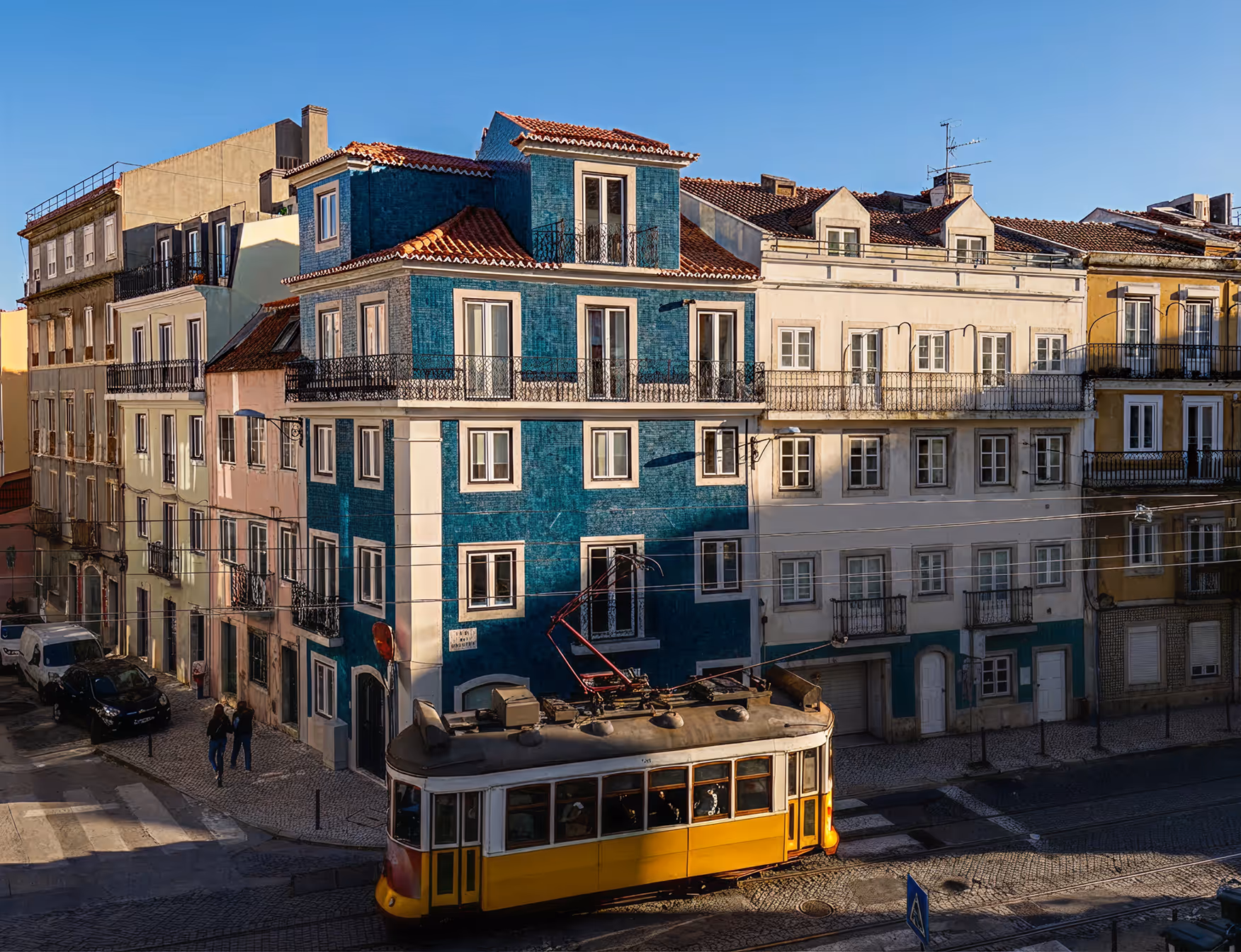 A yellow Lisbon tram passes a sunlit corner of tiled townhouses, including a blue azulejo-fronted building.