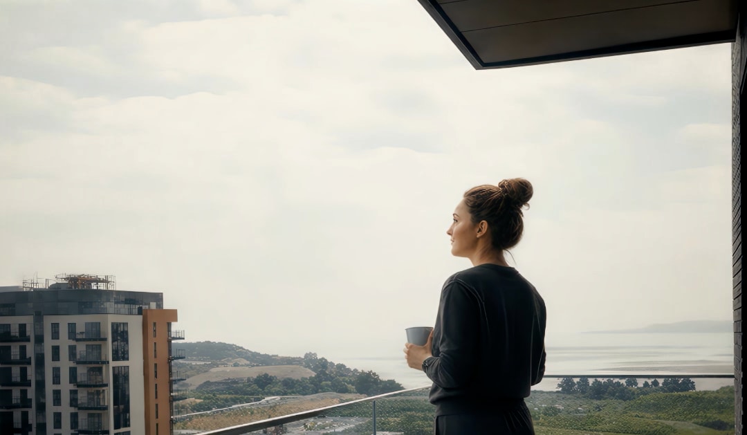 A resident stands on a high-rise apartment balcony with a coffee, looking out over a coastal view beside a neighbouring development under construction.