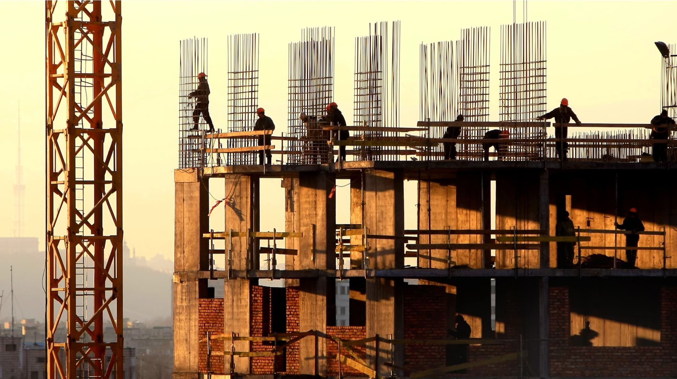 Construction workers on a concrete frame with exposed rebar at golden hour, a tower crane alongside