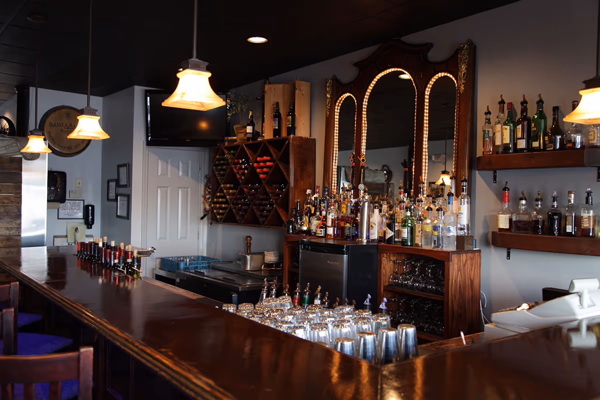 Interior of a bar with wooden countertop, hanging pendant lights, wine bottles on racks, and various liquor bottles displayed behind the bar.