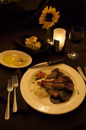 Dinner plate with grilled lamb chops, mashed potatoes, and vegetables next to bread rolls, dipping sauce, glass of red wine, and a sunflower with candle on a dark table.