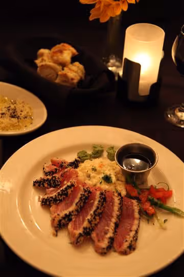 Plate with sesame-crusted seared tuna slices, a creamy side, vegetables, and a dipping sauce in a metal cup, with bread, a candle, and wine glass in the background.