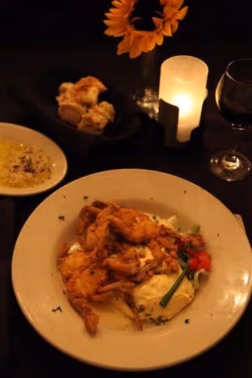 Plate of fried soft-shell crab with mashed potatoes and garnish, next to bread, grated cheese, wine glass, lit candle, and sunflower in a vase.