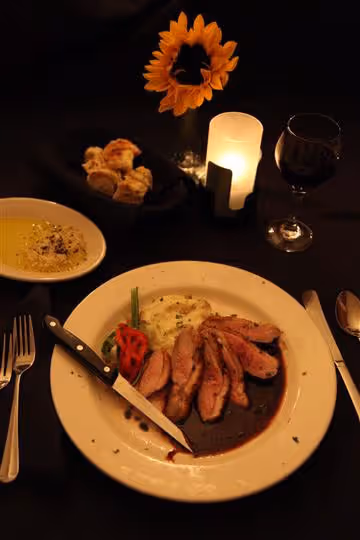 Plate with sliced meat in sauce, mashed potatoes, green beans, and a pepper, accompanied by a knife, fork, and spoon on a black table with a bread basket, soup bowl, candle, wine glass, and a sunflower in a vase.