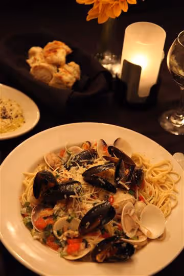 Plate of spaghetti with mussels, clams, diced tomatoes, and grated cheese on a dark table with bread rolls, a glass of white wine, and a lit candle.