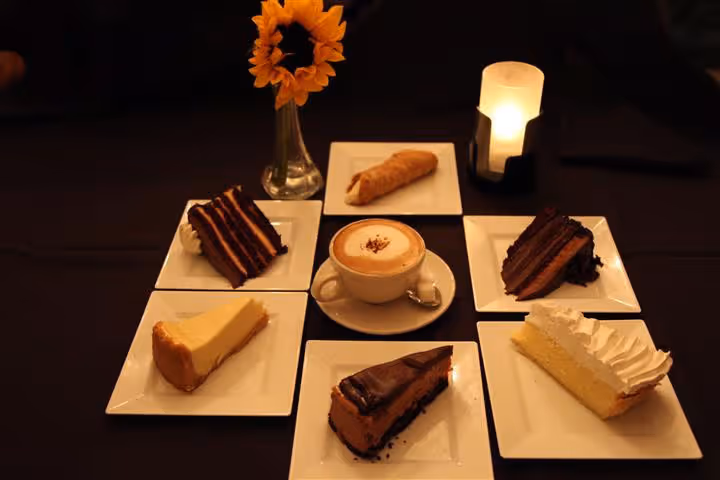 Six dessert plates with various cakes and a cannoli arranged around a cappuccino cup, with a sunflower in a glass vase and a lit candle in the background.