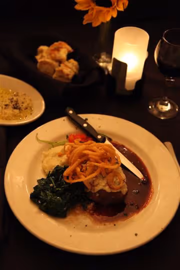 Plated gourmet meal with steak topped with crispy fried onions, mashed potatoes, cooked greens, a bread roll, red wine glass, and lit candle on a dark table.