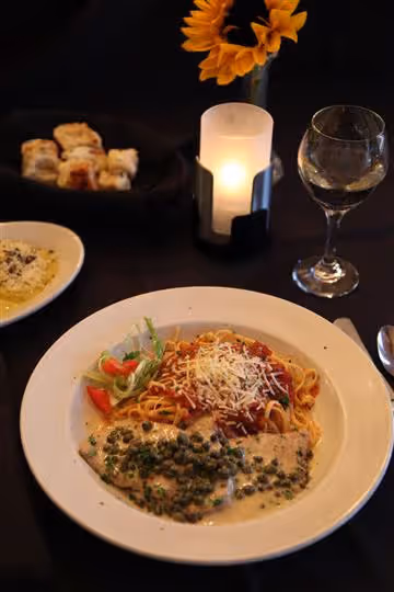 Plate with spaghetti topped with tomato sauce and grated cheese, served with chicken in creamy caper sauce and a small side salad, alongside a glass of white wine, bread, and a lit candle with sunflowers in the background.