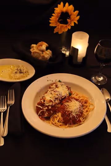 Plate of spaghetti and chicken parmesan with marinara sauce and cheese, bread rolls, dipping sauce, wine glass, sunflower, and candle on a table.