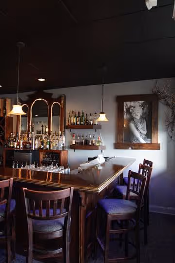 Cozy bar area with wooden stools, a polished wooden counter, shelves with liquor bottles, pendant lights, and a black-and-white portrait on the wall.