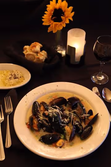 Plate of cooked mussels in broth with grated cheese on a black tablecloth, accompanied by bread rolls, a dish of butter, a glass of red wine, a lit candle, and a sunflower in a vase.