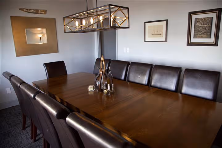 Long wooden dining table with dark leather chairs in a room with framed pictures and a geometric chandelier overhead.