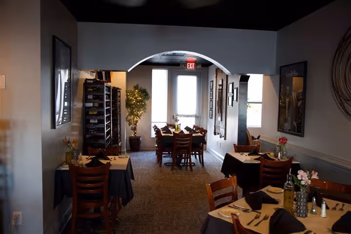Cozy restaurant dining area with wooden chairs, tables set with black napkins and cutlery, and potted plants near a glass door under an archway.