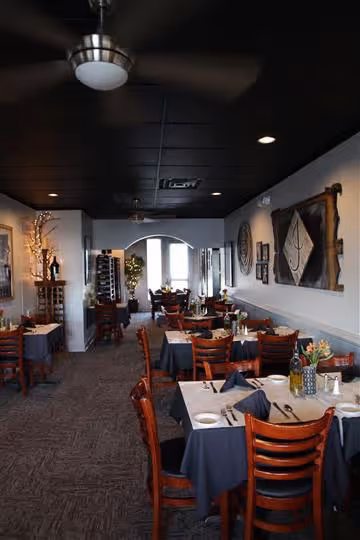 Cozy restaurant interior with wooden chairs and tables set with white plates and dark tablecloths under a black ceiling with ceiling fans.
