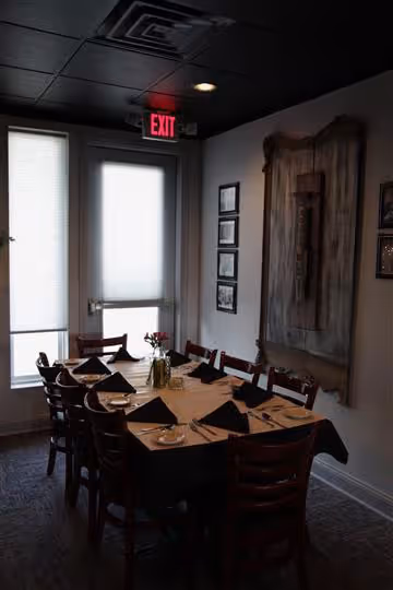 Dimly lit dining room with a table set for eight, featuring black napkins and a flower vase centerpiece, near a door with an illuminated exit sign.