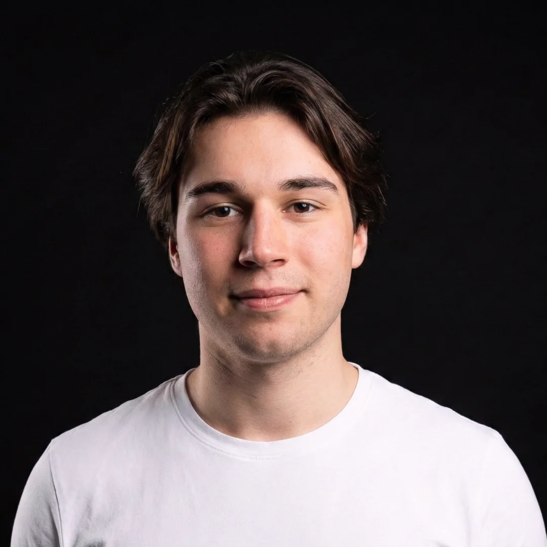Young man with medium-length brown hair wearing a white t-shirt against a black background.