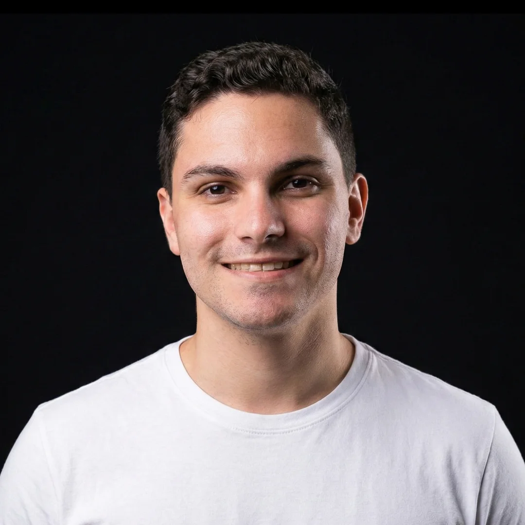 Smiling young man with short dark curly hair wearing a white t-shirt against a black background.