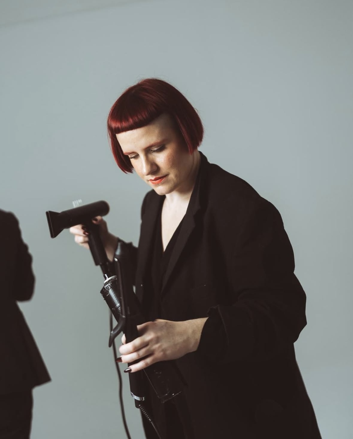 Frau mit rotem Bob-Haarschnitt und schwarzem Mantel hält einen Haartrockner und Lockenstab in der Hand.