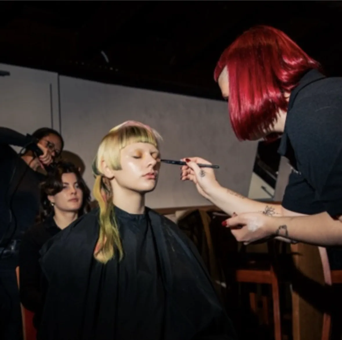 Friseurin mit roten Haaren trägt Kosmetik auf das Gesicht eines Models mit geschlossenen Augen und blondem Haar auf.