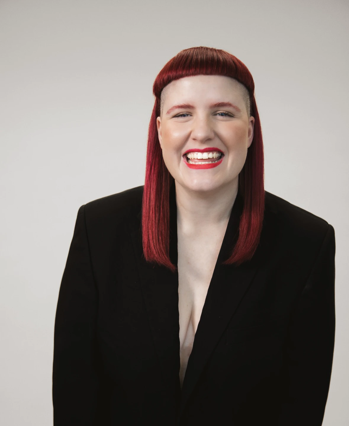 Lachende Person mit roten Haaren, kurzem Pony und seitlich rasiertem Kopf, gekleidet in schwarzes Sakko vor hellem Hintergrund.