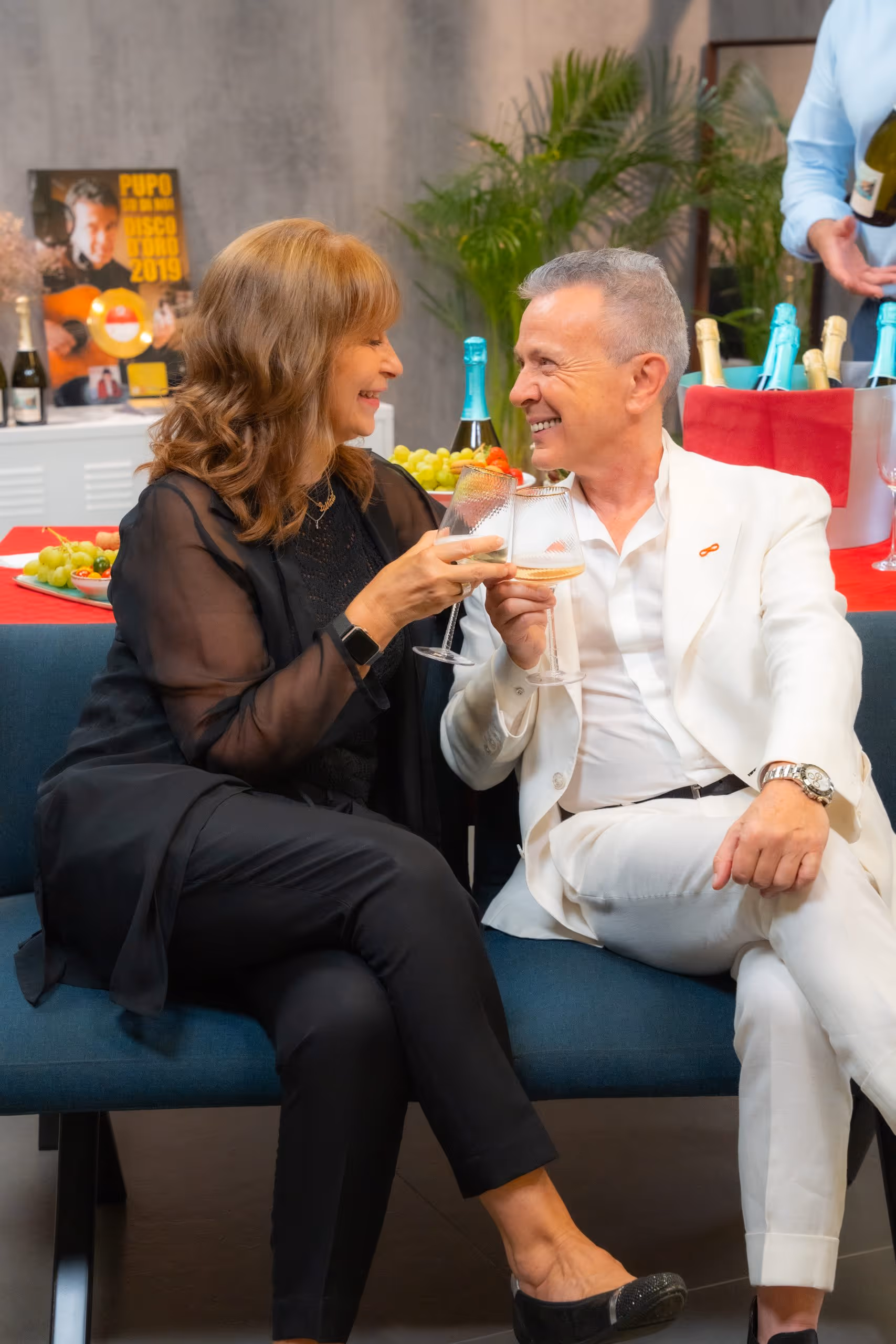A smiling man in a white suit and a woman in black clothes clinking glasses while sitting on a blue bench at a celebration with wine and fruits in the background.
