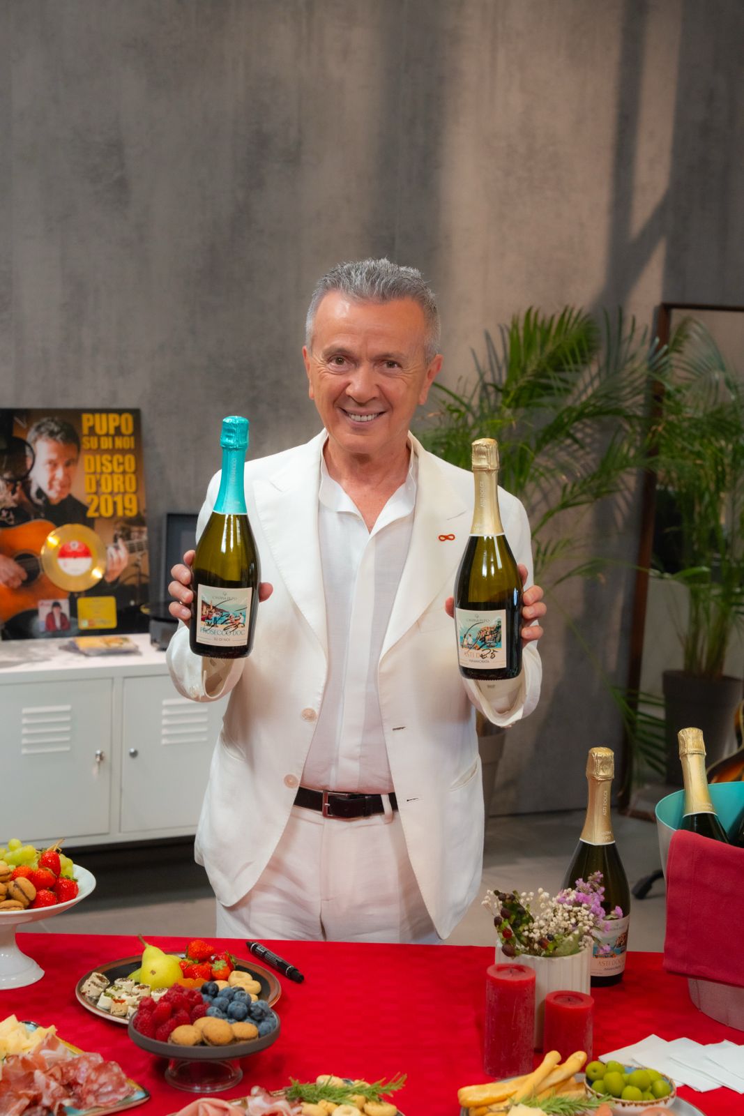 Smiling man in a white suit holding two bottles of sparkling wine in a decorated room with a red tablecloth and assorted snacks.