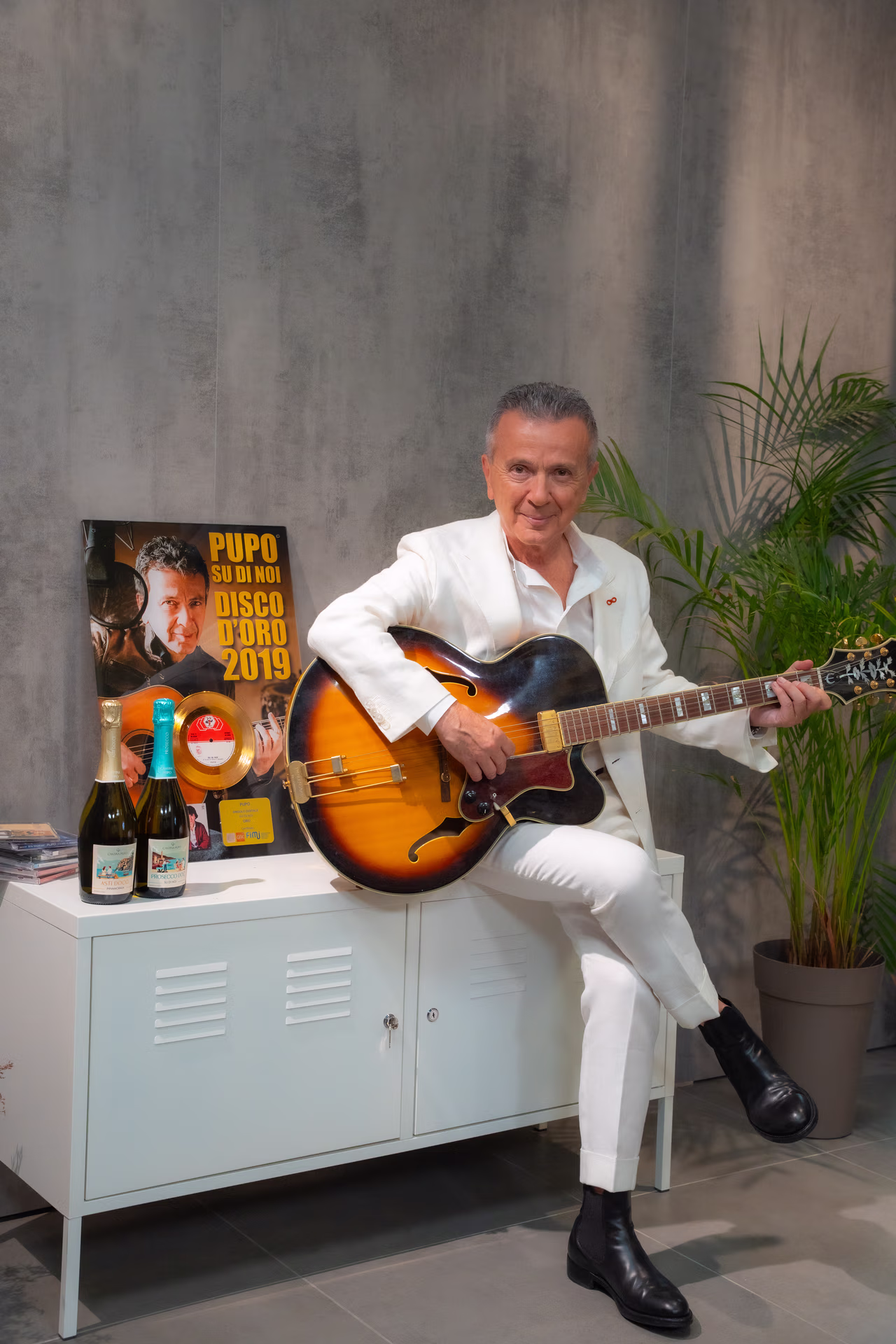Man in a white suit sitting on a white cabinet playing a sunburst electric guitar, with a gold record award and two bottles on the cabinet, and a tall green plant in the background.