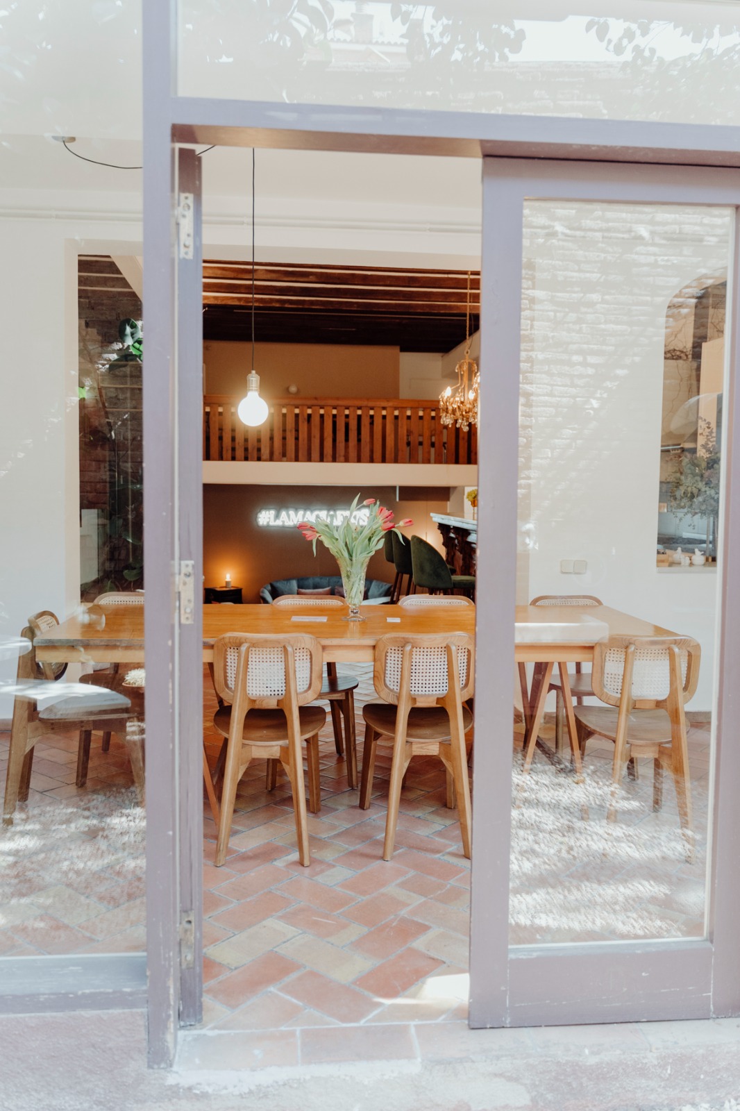 Comedor con mesa de madera larga rodeada de sillas, con jarrón de flores en el centro y ventana corrediza de vidrio, así es nuestro estudio en Barcelona.