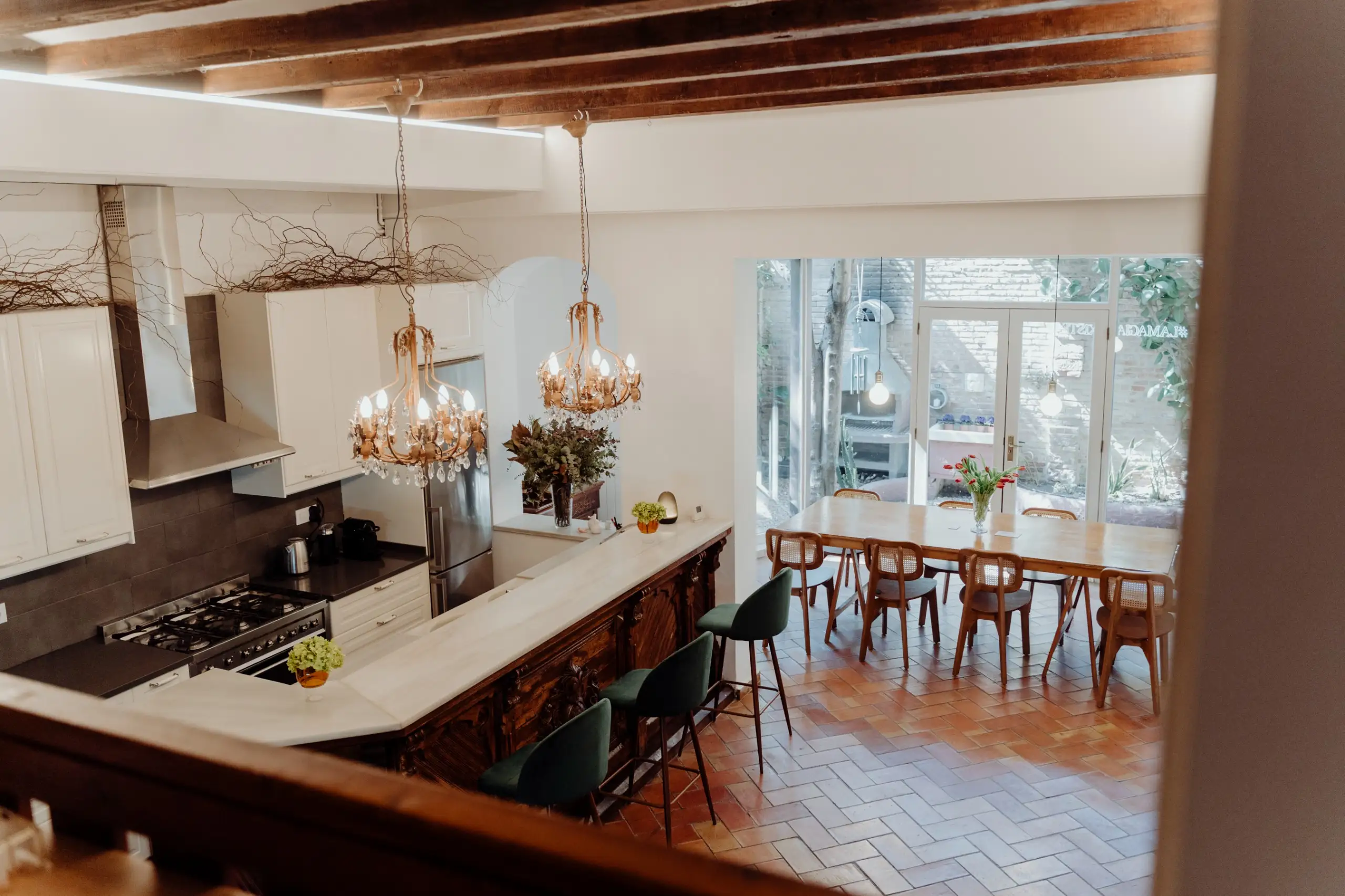 Cocina abierta con barra de mármol, tres sillas altas verdes y mesa de comedor de madera con sillas junto a una ventana grande que da al jardín, así es el estudio de INOCUO The Sign en Barcelona.