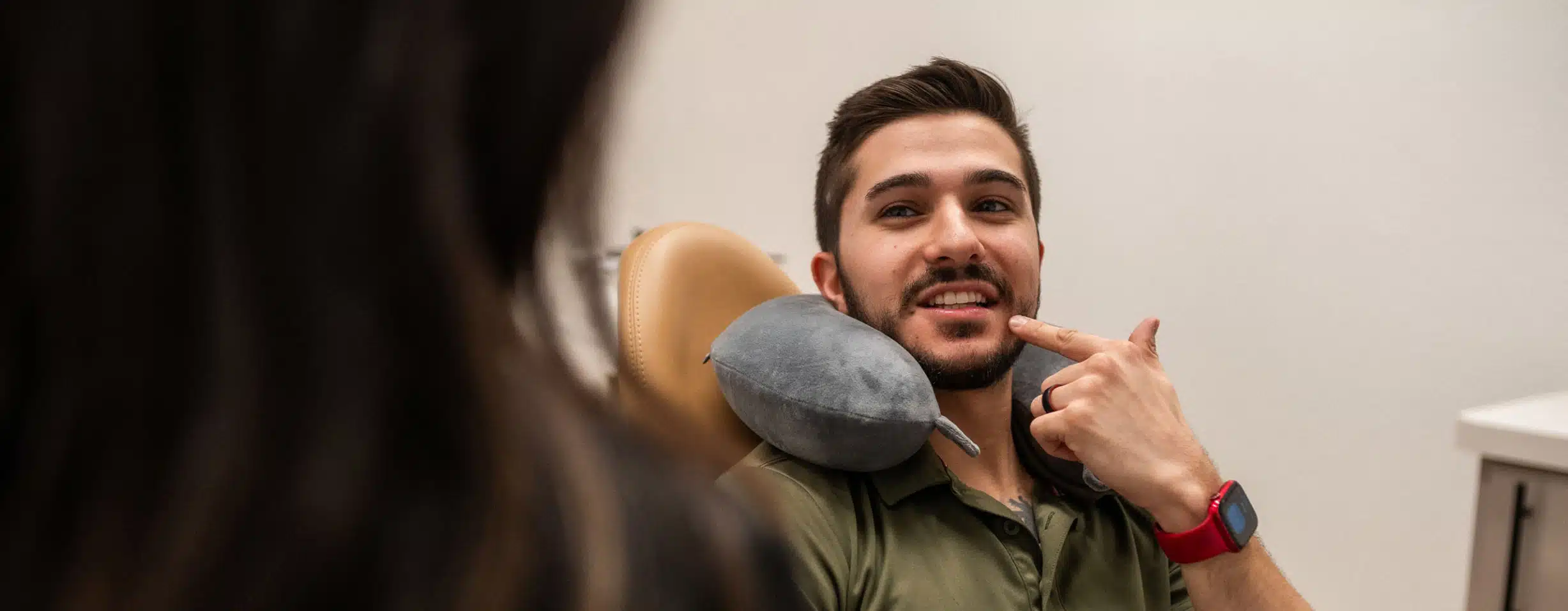 Man sitting in a chair with a neck pillow pointing to his teeth, smiling and talking to someone.