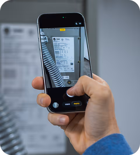 Hand holding smartphone capturing photo of a Trane equipment label with technical details.