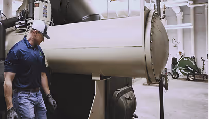 Man in gloves and cap inspecting large industrial ventilation or HVAC duct in a utility room.