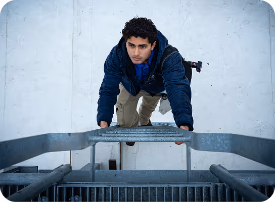 Young man in a blue jacket climbing a metal ladder attached to a white wall, viewed from above.