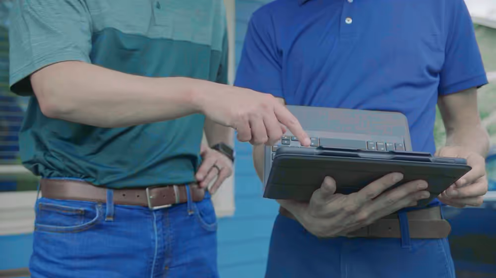 Two men using and pointing at a tablet device outside a building with blue siding.