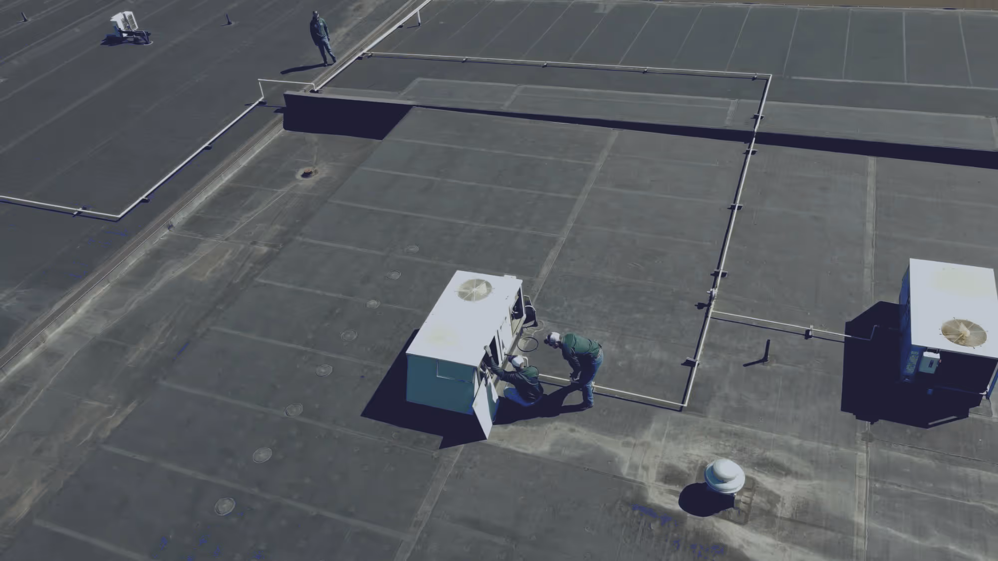 Two workers in helmets repairing rooftop HVAC unit with another person standing nearby.