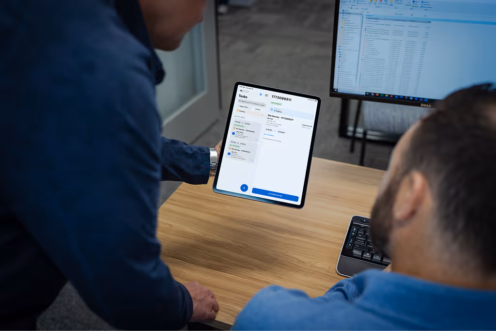 Two men looking at a tablet displaying a tasks app, sitting at a desk with a computer monitor and keyboard.