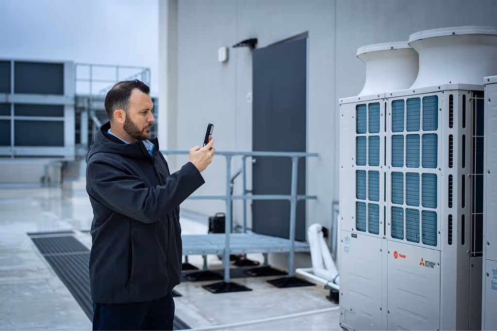 Technician inspecting HVAC units on rooftop, using a smartphone for assessment.