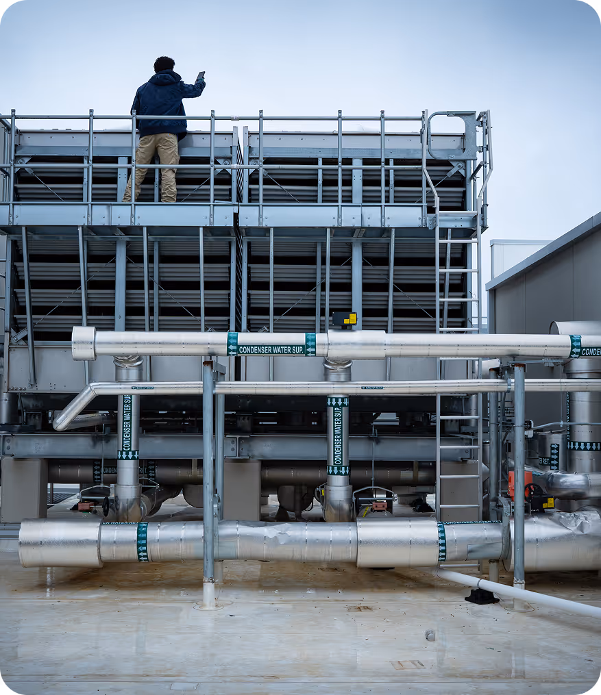 Technician in jacket and khakis inspecting industrial chiller system on rooftop with labeled condenser pipes.