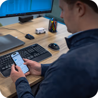 Person holding smartphone with app interface in front of a computer keyboard and mouse on a desk.