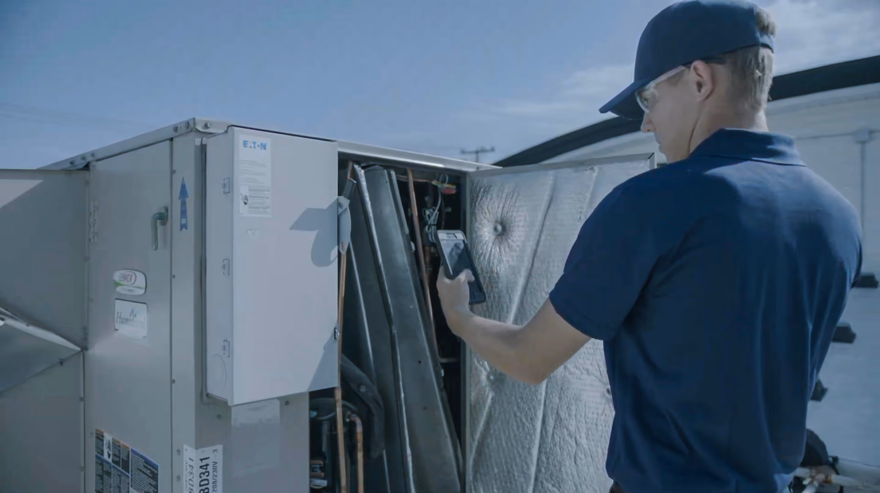Technician in blue uniform using a smartphone to inspect the inside of an outdoor HVAC unit.