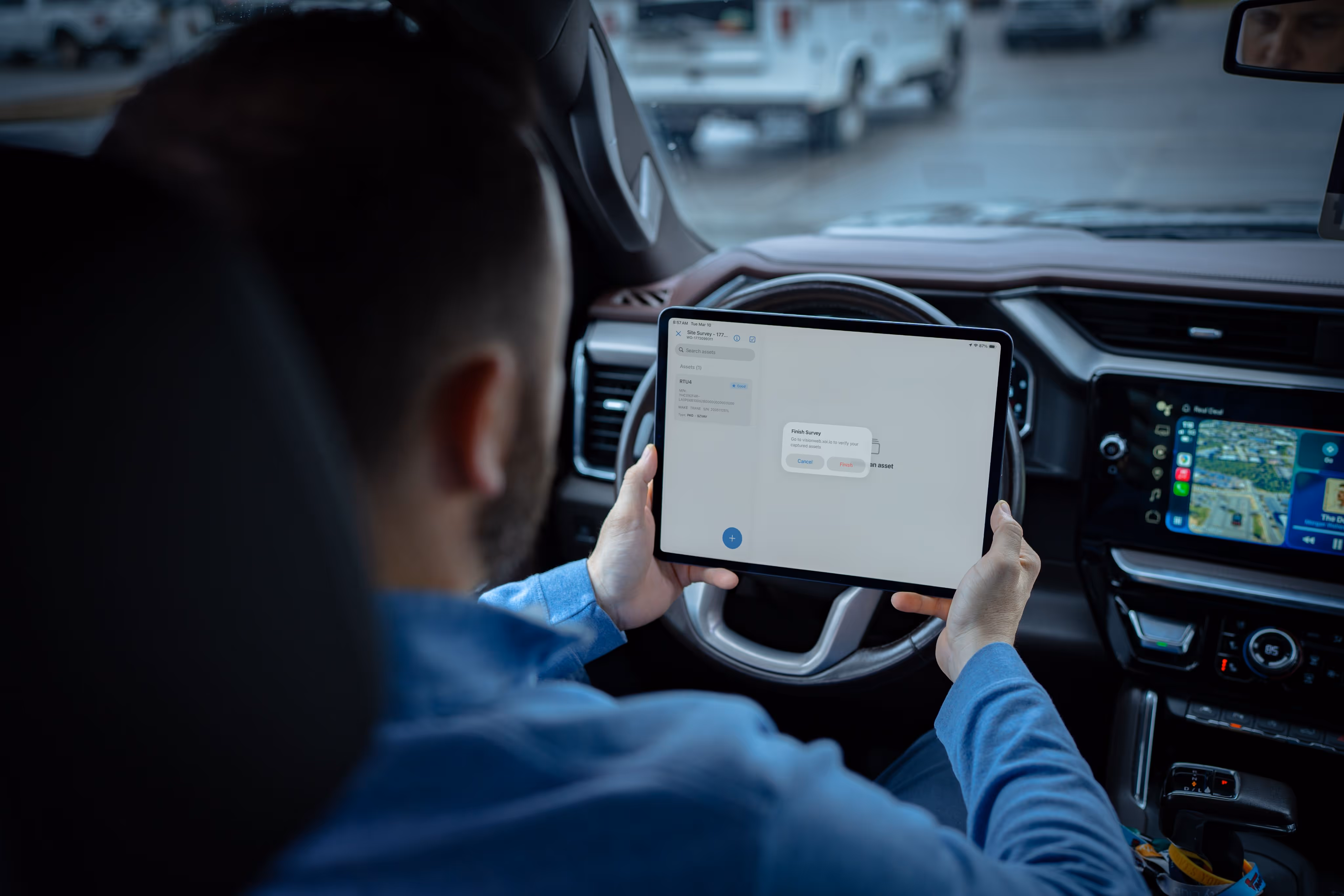 Man in blue shirt holding a tablet inside a vehicle with a navigation screen on the dashboard.