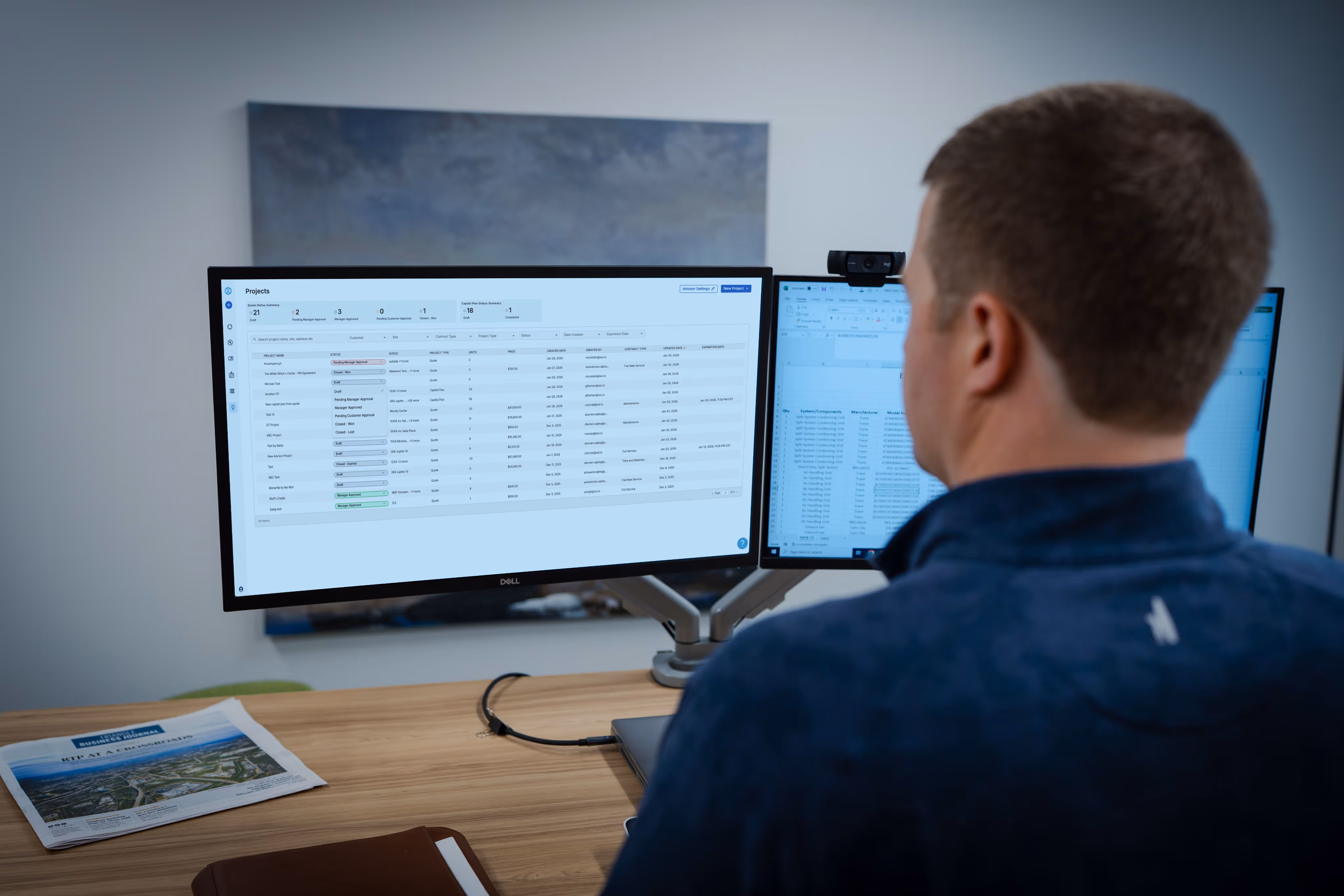 Man working at dual monitors displaying project management and spreadsheet data at a desk with papers.
