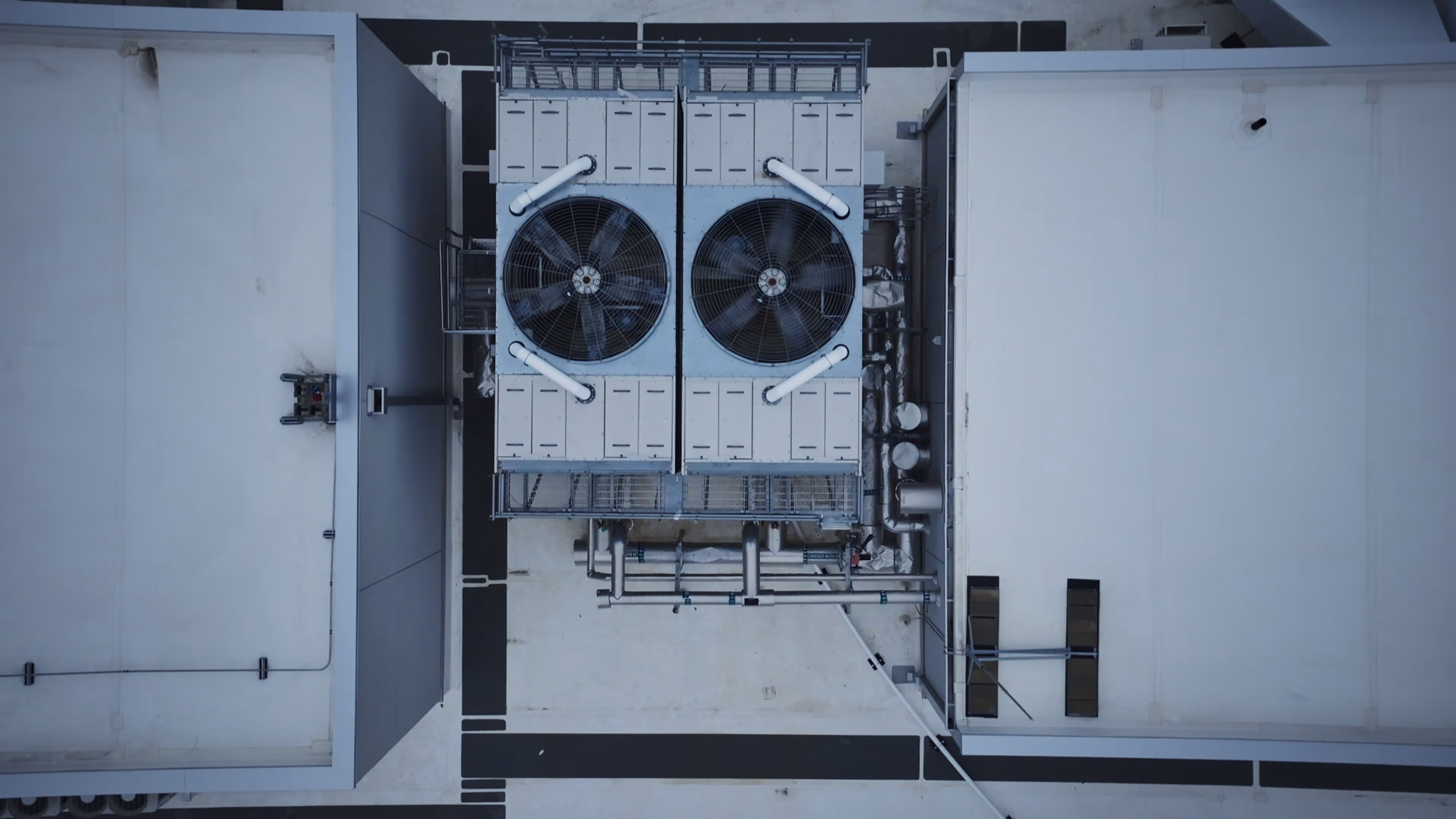 Aerial view of two large industrial cooling fans on a rooftop surrounded by pipes and building structures.