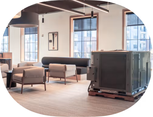 Modern office lounge with beige chairs, a black couch, large windows, and a large metal machine on a pallet.