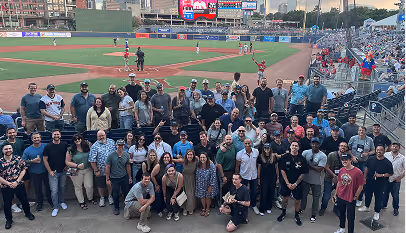 Large group of people posing and smiling at a baseball stadium with the field and players visible behind them.