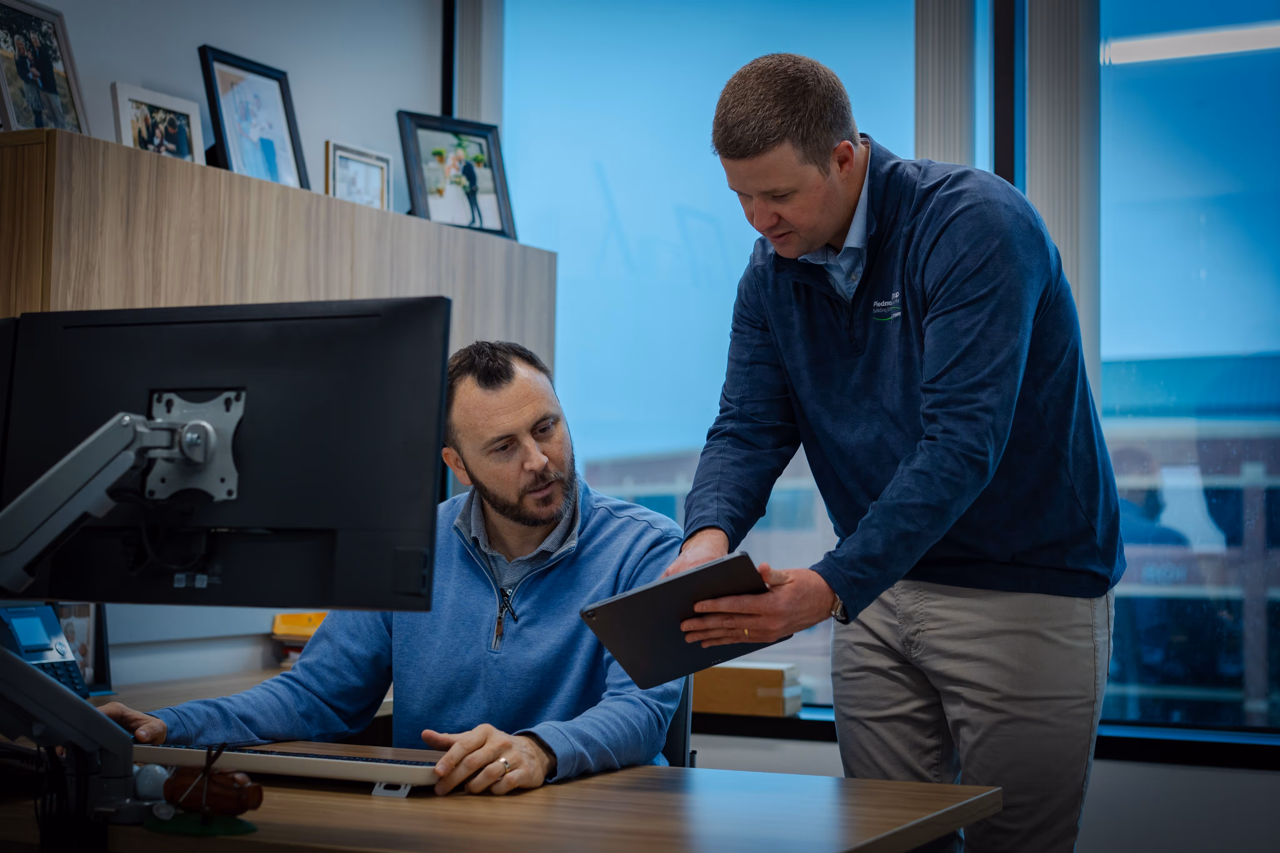 Two men in an office, one seated at desk with computer, the other showing him a tablet.