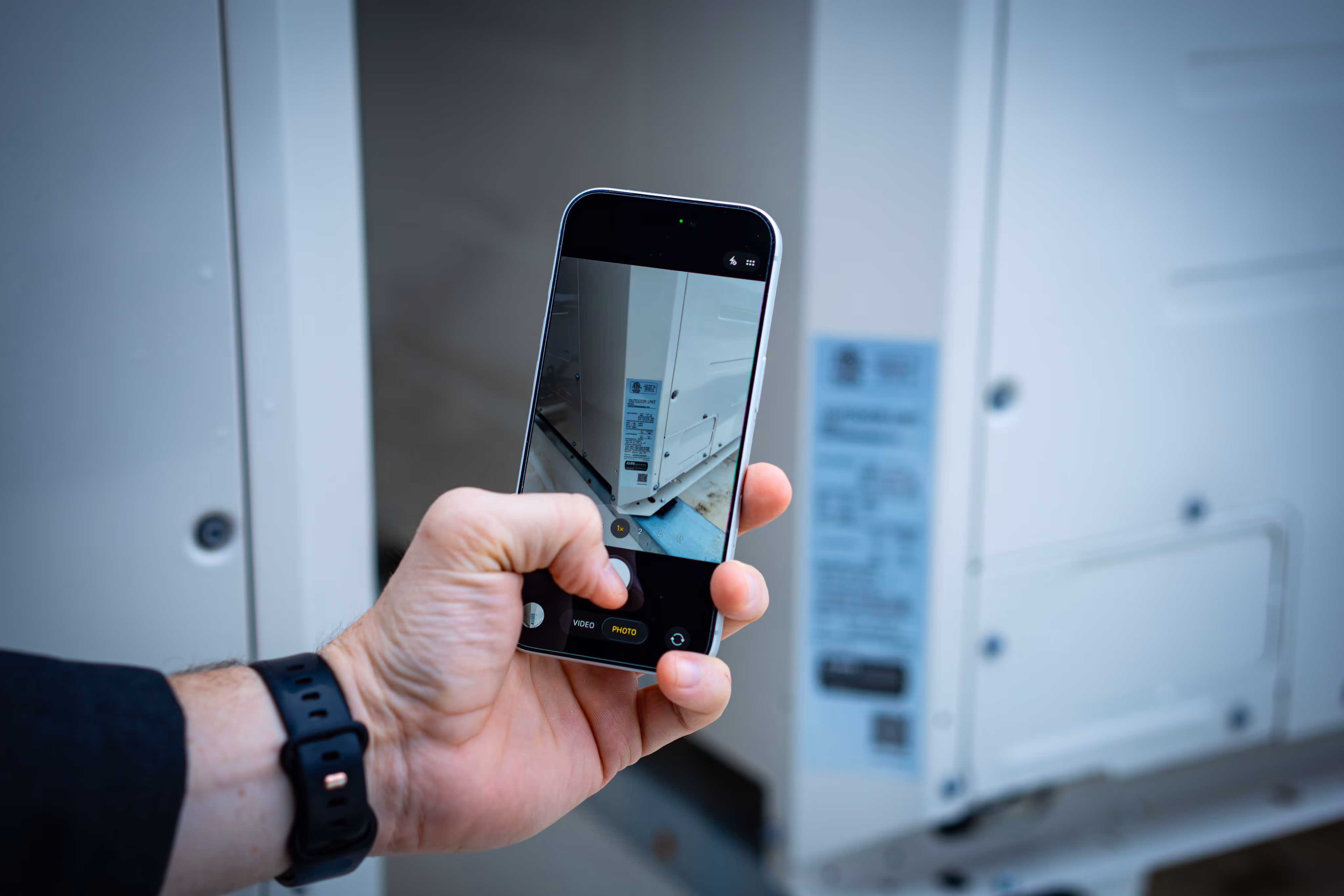Hand holding smartphone taking a close-up photo of a white industrial unit with a label.