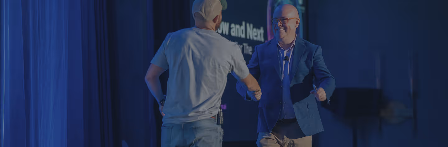 Two men shaking hands on a dimly lit stage with blue curtains and a screen in the background.