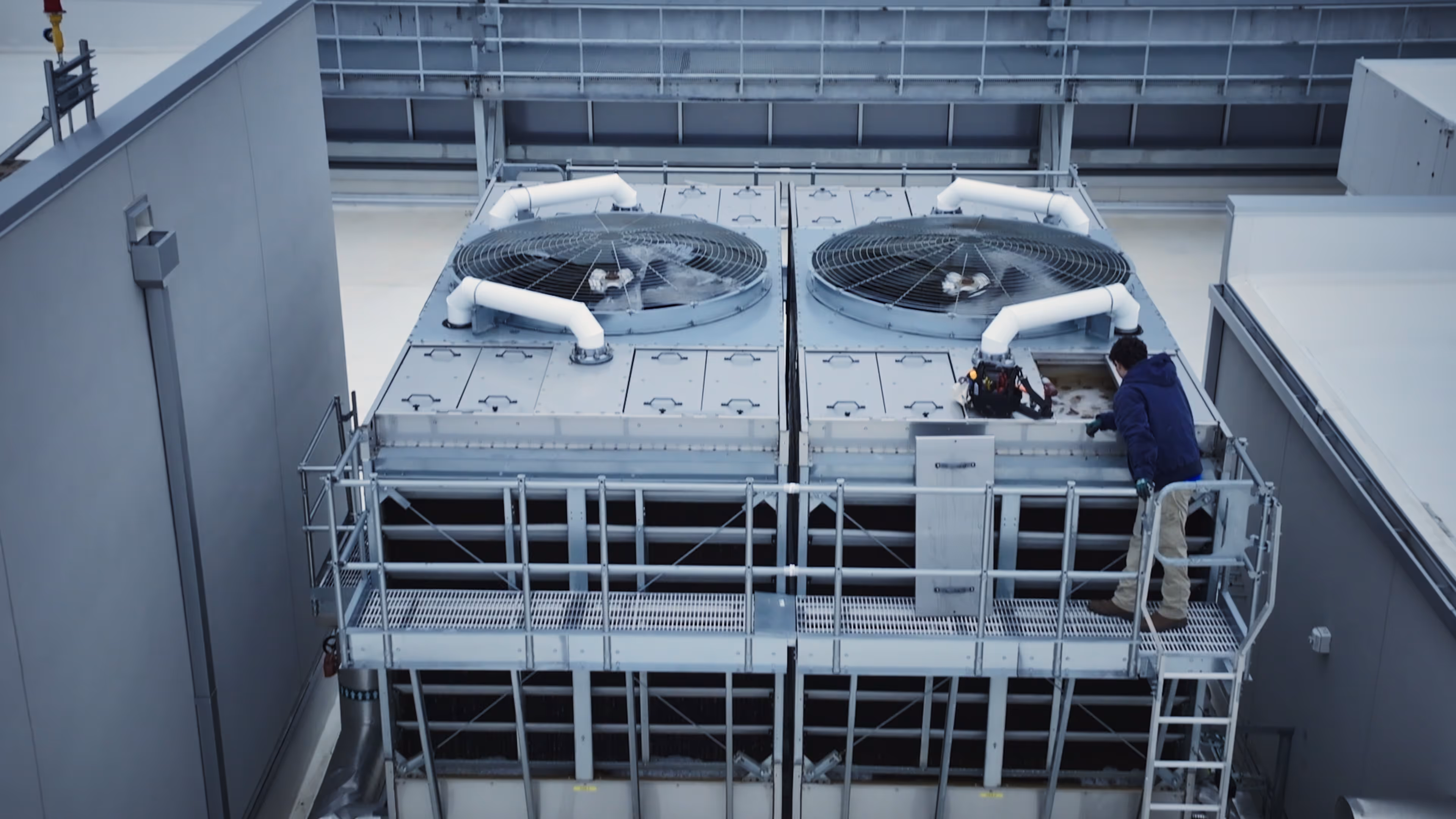 Technician inspecting one of two large rooftop HVAC units with fans and pipes.