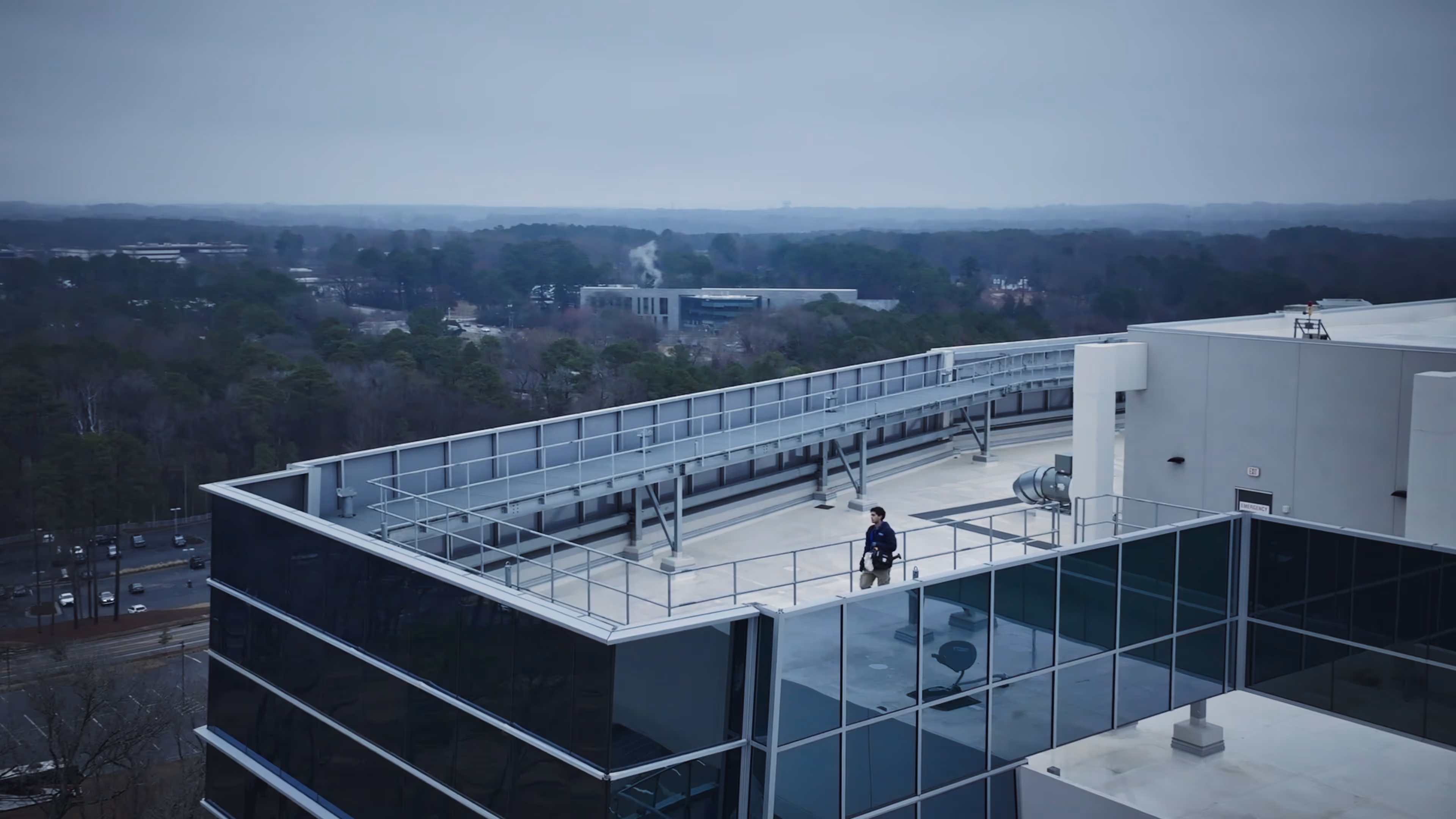 Man walking on rooftop of modern building with trees and distant buildings under cloudy sky.